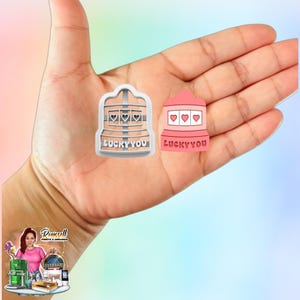May include: Two slot machine-shaped cookie cutters. One is white, the other is pink. Both have three heart symbols and the text "LUCKY YOU". The cookie cutters are held in a hand.