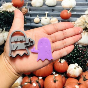 May include: A gray ghost-shaped cookie cutter and a purple ghost-shaped object are displayed. The cookie cutter has a handle and a ghost outline. The purple ghost has a thumbs-up gesture. The background includes pumpkins and gourds.