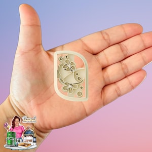 May include: A beige, rectangular cookie cutter with a rounded corner, featuring a butterfly and floral design. The cutter is held in a hand against a gradient pink and blue background. The design includes a butterfly, flowers, and small circles.