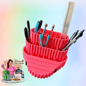 May include: A bright pink, tiered desk organizer with multiple compartments. It holds various tools, including pens, scissors, and craft tools. The organizer has a unique, wavy design and is set against a colorful, gradient background.