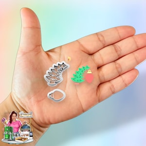 May include: A hand holding four Christmas-themed cookie cutters. The cutters include a Christmas tree, a Christmas tree with a red ornament, a light bulb, and a small star. The cutters are made of silver metal.