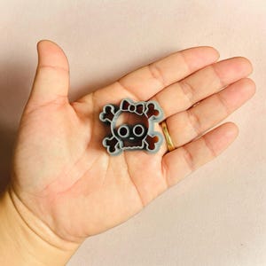 May include: A gray plastic cookie cutter shaped like a skull with a bow on top. The skull has two eye holes and a small mouth.