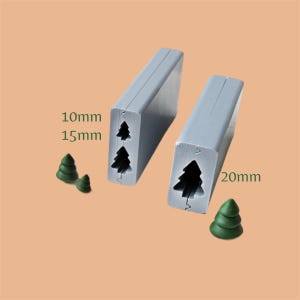 May include: Two gray rectangular metal molds with Christmas tree cutouts, measuring 10mm, 15mm, and 20mm. Two small green Christmas tree-shaped decorations are also present. The background is a solid light orange color.