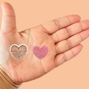 May include: Two heart-shaped yarn balls, one white and one pink, are shown on a person's open palm. The yarn balls have a textured, woven pattern.