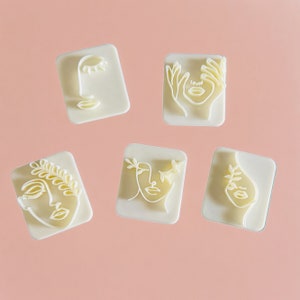 May include: Six white plastic stamps with different line art designs of faces. The stamps are on a light pink background.