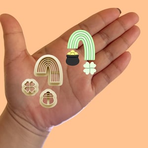 May include: A set of four cookie cutters featuring a rainbow, a pot of gold, and two four-leaf clovers. The cutters are made of a light brown plastic and are perfect for making St. Patrick's Day cookies.