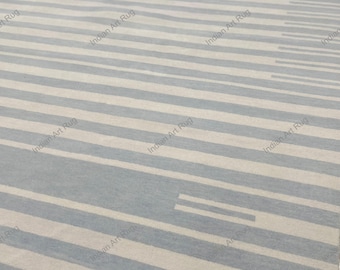 May include: A flat-woven rug featuring a repeating pattern of horizontal stripes in alternating shades of light gray and off-white. The surface displays a consistent textile texture with the text Indian Art Rug overlaid repeatedly.