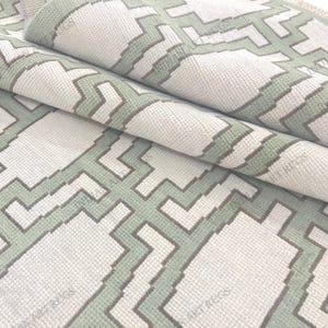 May include: A rolled-up area rug with a geometric pattern. The rug features a repeating design of light green and gray shapes on a white background. The texture of the rug is visible, suggesting a soft, woven material. The rug is folded over itself.