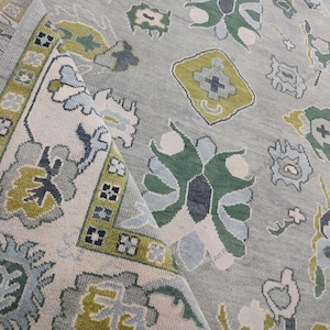 May include: A folded, light gray rug with a detailed floral pattern. The design incorporates shades of green, blue, and cream. The border features geometric shapes and a contrasting color scheme, adding visual interest to the textile.