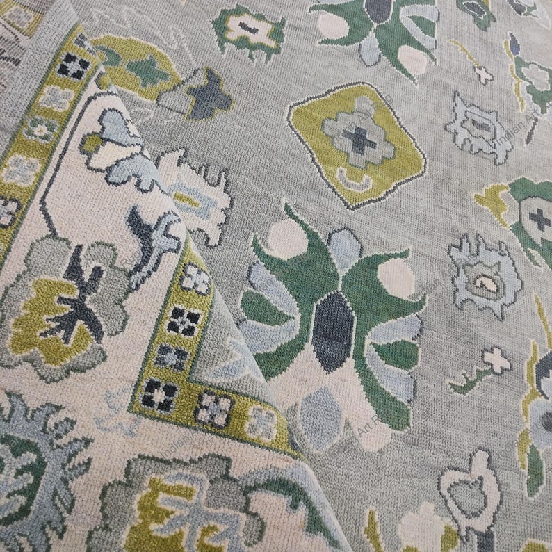 May include: A folded, light gray rug with a detailed floral pattern. The design incorporates shades of green, blue, and cream. The border features geometric shapes and a contrasting color scheme, adding visual interest to the textile.