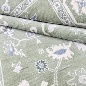 May include: A sage green area rug featuring a repeating floral and geometric pattern in white and soft blue. The textile displays a dense, tufted pile with intricate, symmetrical motifs and the repeated watermark text Indian Art Rug.