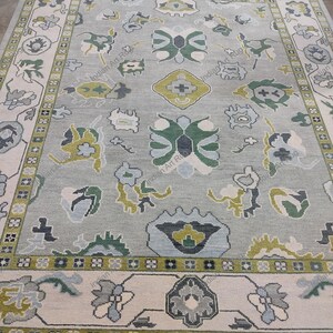 May include: A rectangular area rug with a light gray background and a detailed floral pattern in shades of green, blue, and cream. The rug has a cream-colored border with a repeating floral design and fringe along the edges.