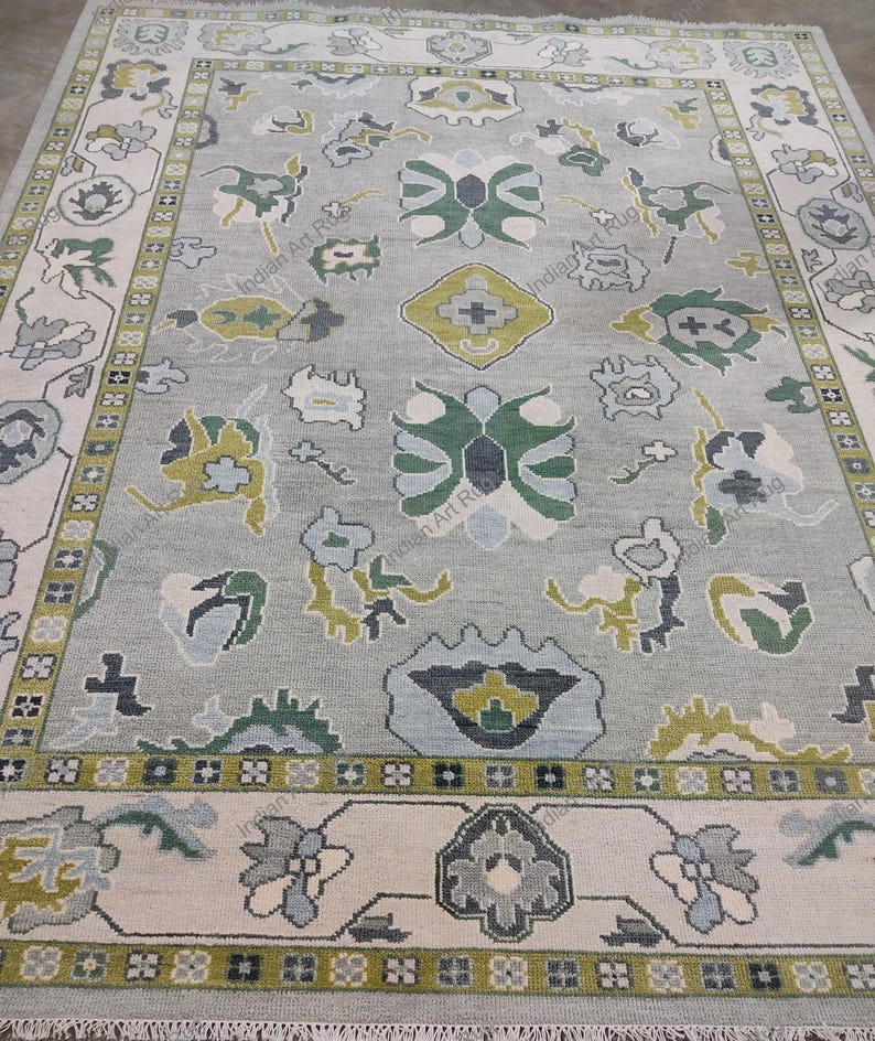 May include: A rectangular area rug with a light gray background and a detailed floral pattern in shades of green, blue, and cream. The rug has a cream-colored border with a repeating floral design and fringe along the edges.