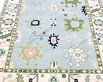 Puede incluir: Una alfombra rectangular con un campo central azul claro decorado con motivos florales abstractos dispersos en tonos verde, rosa, blanco y azul oscuro, enmarcada por un borde ancho color crema con patrones geométricos verdes.