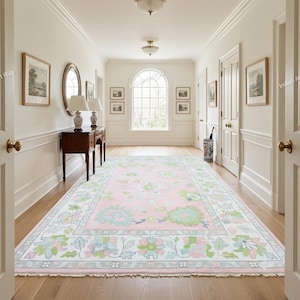 May include: A rectangular area rug featuring a soft pink background with a repeating floral pattern in shades of light green, white, and pale blue. The rug has a wide border with intricate botanical motifs and short white fringe on both ends.
