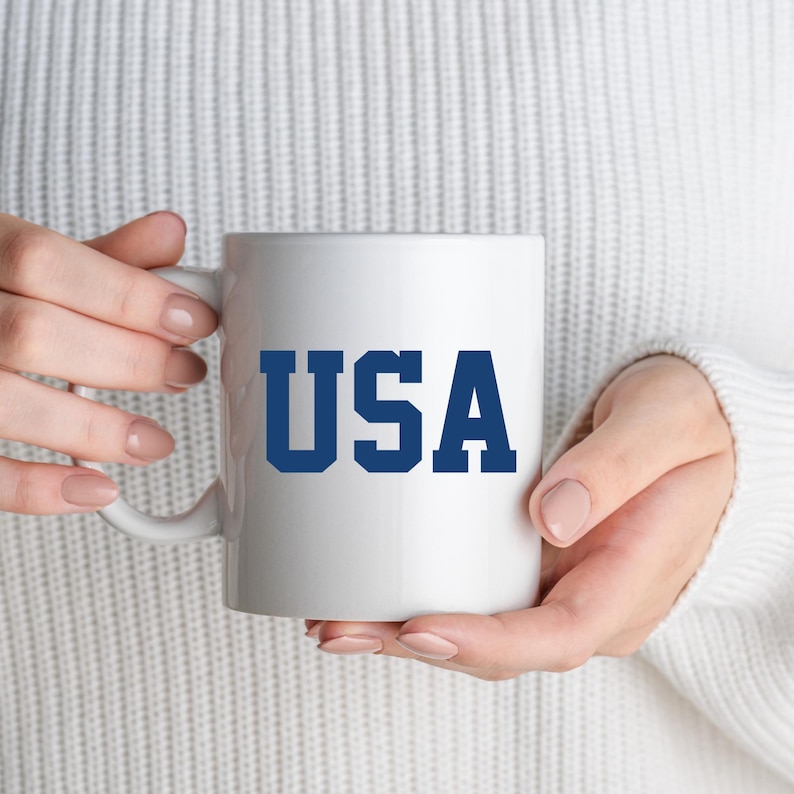 May include: White ceramic mug with the word "USA" in bold, navy blue letters. The mug is held by a person wearing a white sweater. The mug has a standard handle and a cylindrical shape.