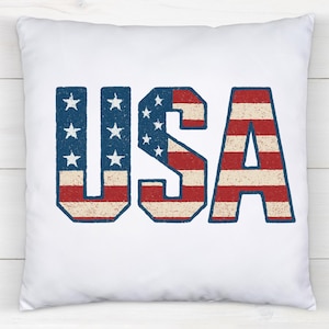 May include: White square pillow with the word "USA" in distressed red, white, and blue lettering. The letters "U" and "S" feature blue backgrounds with white stars, while the "A" has red and white stripes.