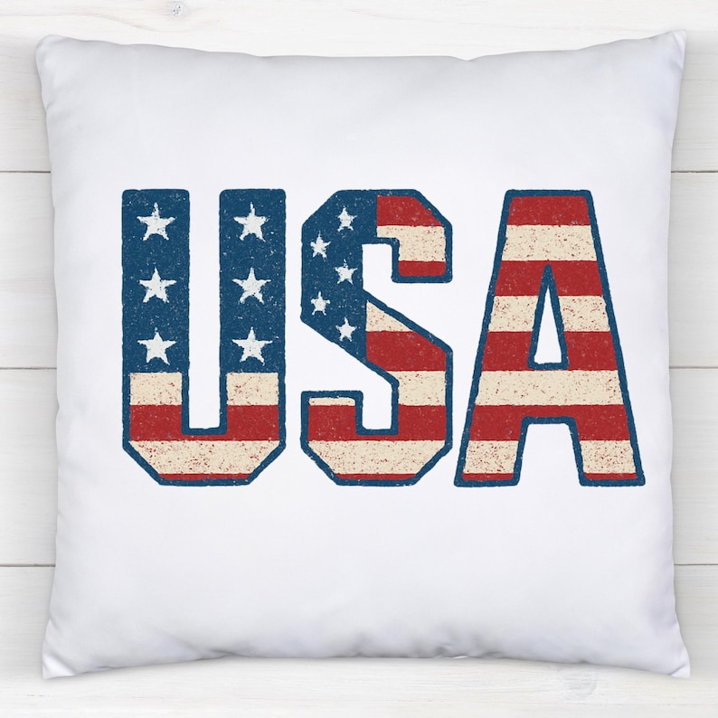 May include: White square pillow with the word "USA" in distressed red, white, and blue lettering. The letters "U" and "S" feature blue backgrounds with white stars, while the "A" has red and white stripes.