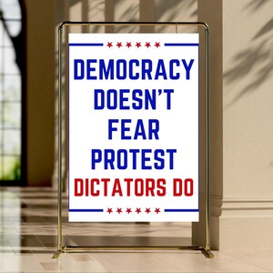 May include: A white sign with the text "DEMOCRACY DOESN'T FEAR PROTEST DICTATORS DO" in blue and red, displayed on a gold metal stand. The sign has blue and red star accents.