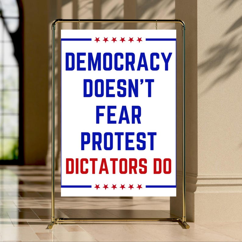 May include: A white sign with the text "DEMOCRACY DOESN'T FEAR PROTEST DICTATORS DO" in blue and red, displayed on a gold metal stand. The sign has blue and red star accents.