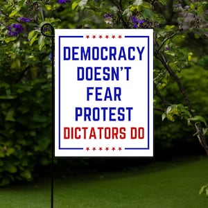 May include: A rectangular garden flag with a white background and blue border. The flag features the text "DEMOCRACY DOESN'T FEAR PROTEST DICTATORS DO" in blue and red, with star accents. The flag is displayed outdoors.