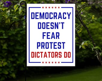 May include: A rectangular garden flag with a white background and blue border. The flag features the text "DEMOCRACY DOESN'T FEAR PROTEST DICTATORS DO" in blue and red, with star accents. The flag is displayed outdoors.