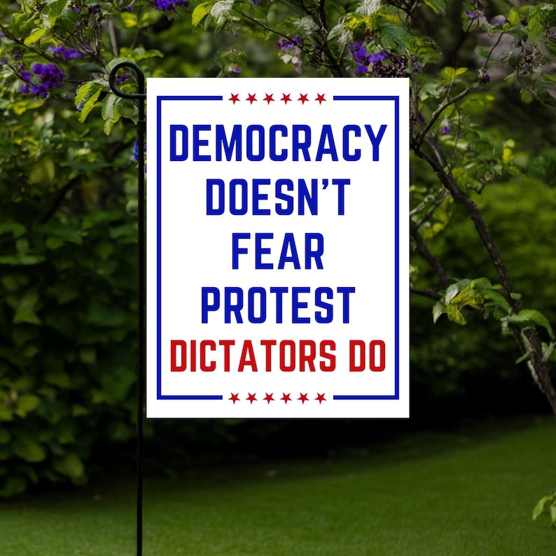 May include: A rectangular garden flag with a white background and blue border. The flag features the text "DEMOCRACY DOESN'T FEAR PROTEST DICTATORS DO" in blue and red, with star accents. The flag is displayed outdoors.