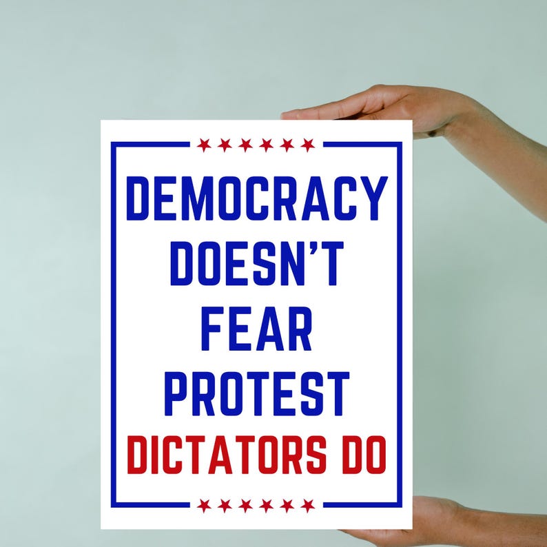 May include: A white poster with a blue border and the text "DEMOCRACY DOESN'T FEAR PROTEST DICTATORS DO" in blue and red. The top and bottom of the poster have a row of red stars. The poster is held up by a person.