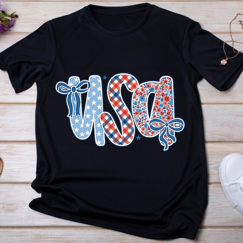 May include: Black t-shirt with the word "USA" in a patriotic design. The letters are decorated with stars, gingham, and floral patterns in red, white, and blue. A bow embellishes the letters. The shirt is on a wooden surface.