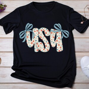 May include: A navy blue t-shirt with the word "USA" in a floral pattern. The letters are decorated with light blue bows. The floral pattern includes red and blue flowers on a cream background.