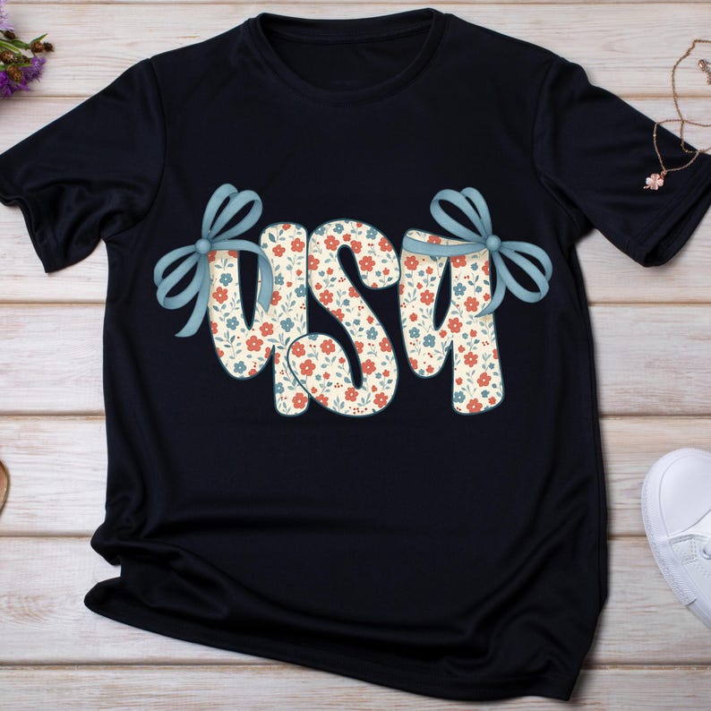 May include: A navy blue t-shirt with the word "USA" in a floral pattern. The letters are decorated with light blue bows. The floral pattern includes red and blue flowers on a cream background.