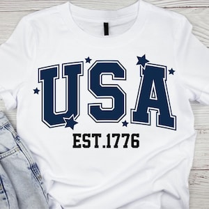 May include: White t-shirt with the word "USA" in navy blue block letters, accented with stars. Below the text is "EST.1776" in a black, bold font. The shirt is made of a soft material.