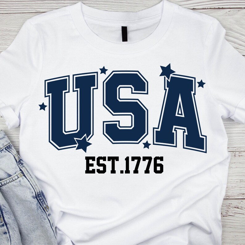 May include: White t-shirt with the word "USA" in navy blue block letters, accented with stars. Below the text is "EST.1776" in a black, bold font. The shirt is made of a soft material.