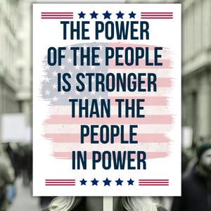 May include: A white sign with the text "THE POWER OF THE PEOPLE IS STRONGER THAN THE PEOPLE IN POWER" in dark blue. The sign features a faded American flag design in the background, with red and blue accents.