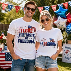 May include: Two white t-shirts featuring patriotic graphics. The left shirt displays the text I LOVE THE TASTE OF FREEDOM with an American flag and fireworks. The right shirt features the word FREEDOM with an American flag and fireworks.