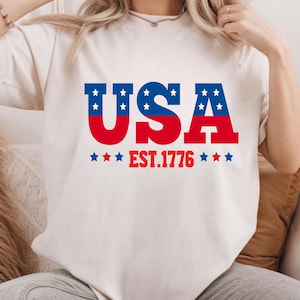 May include: Cream-colored t-shirt with a patriotic design. The word "USA" is printed in red and blue, with white stars in the letters. Below the word "USA" is the text "EST.1776" in red.