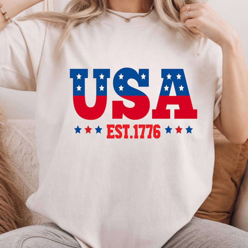May include: Cream-colored t-shirt with a patriotic design. The word "USA" is printed in red and blue, with white stars in the letters. Below the word "USA" is the text "EST.1776" in red.