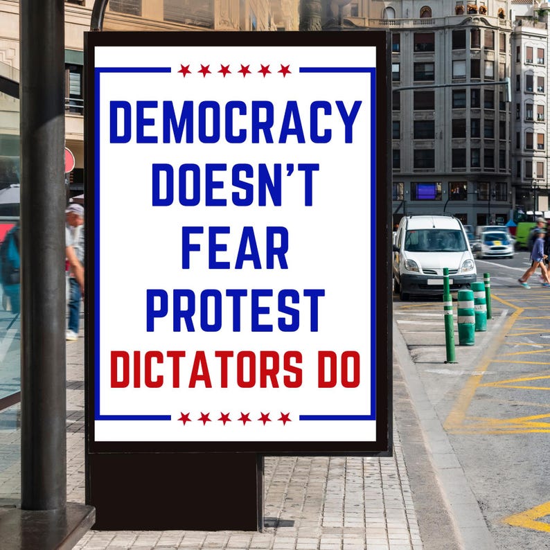 May include: An outdoor advertisement with the text "DEMOCRACY DOESN'T FEAR PROTEST DICTATORS DO" in blue and red. The sign is framed in black and has a white background. The background shows a city street with cars and pedestrians.
