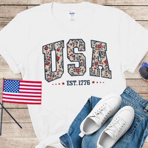 May include: White t-shirt with the word "USA" in a floral pattern, "EST. 1776" below. The image includes a small American flag, sunglasses, white sneakers, and blue jeans. Patriotic theme.