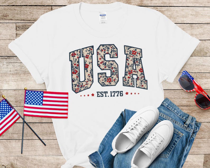 May include: White t-shirt with the word "USA" in a floral pattern, "EST. 1776" below. The image includes a small American flag, sunglasses, white sneakers, and blue jeans. Patriotic theme.
