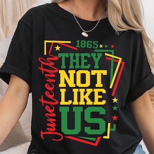 May include: Black t-shirt featuring a vibrant graphic. The design includes the word "Juneteenth" in red, and the phrase "THEY NOT LIKE US" in green and yellow, with the year "1865" above. The shirt has a crew neck and short sleeves.