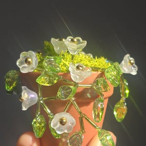 Pode incluir: Um pequeno vaso de terracota cheio de musgo verde e adornado com flores e folhas feitas à mão. As flores são brancas com centros dourados, e as folhas são de um verde vibrante. Um charmoso elemento decorativo floral.
