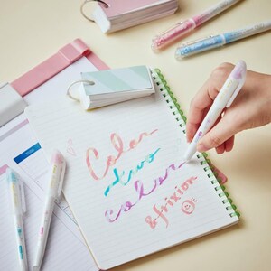 Peut inclure: Une main tenant un stylo effa&ccedil;able blanc avec un motif floral rose et bleu. Le stylo est utilis&eacute; pour &eacute;crire les mots "Color, two colors, frixion" sur un carnet blanc avec une reliure spirale verte.