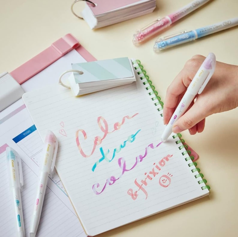 Peut inclure: Une main tenant un stylo effa&ccedil;able blanc avec un motif floral rose et bleu. Le stylo est utilis&eacute; pour &eacute;crire les mots "Color, two colors, frixion" sur un carnet blanc avec une reliure spirale verte.