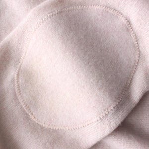 Puede incluir: Un suéter de punto rosa claro que presenta un parche ovalado a juego en el codo. El parche está sujeto firmemente a la manga con costuras tonales visibles alrededor del perímetro, integrándose perfectamente en la tela suave y texturizada.