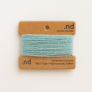 May include: A spool of light blue yarn is wrapped around a brown cardboard tag. The tag has the text ".nd SECOND CASHMERE" at the top and ".nd Reclaimed Cashmere 10m // 2-ply // Approximately 2/28nm" at the bottom. The yarn is a soft, muted blue.