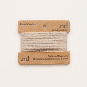 May include: A spool of light beige reclaimed cashmere yarn wrapped around a rectangular brown cardboard card. The card features the text .nd, Made in Scotland, Second Cashmere, 10m // 2-ply // Approximately 2/28nm printed in dark brown.