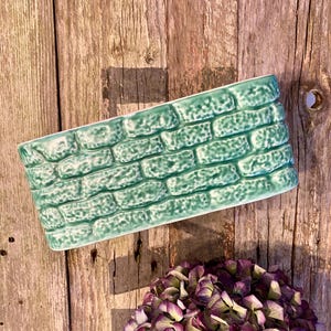May include: A rectangular, light green ceramic planter with a brick pattern. The planter is set against a rustic wooden background, with a cluster of purple and green hydrangeas in the foreground. The planter is approximately 25 cm long.