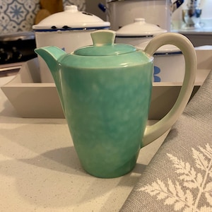 May include: A turquoise ceramic teapot with a white handle and lid. The teapot is on a white countertop, with a gray and white patterned napkin in the foreground. Other white enamelware pots are in the background.