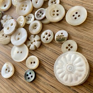 May include: A selection of off-white buttons in various shapes and sizes, including round and flower-shaped designs. Some have four holes, others two. The buttons are displayed on a light brown wooden surface.